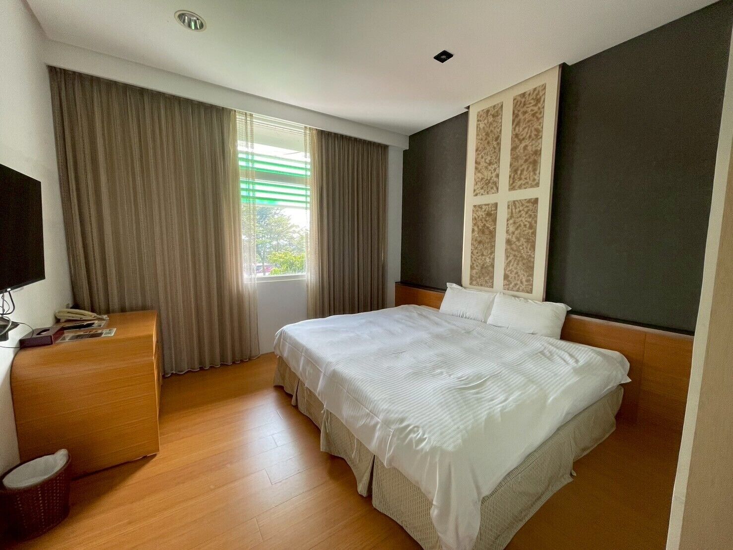 A hotel room with a large bed, a wooden dresser, a flat-screen TV, and a window with curtains.