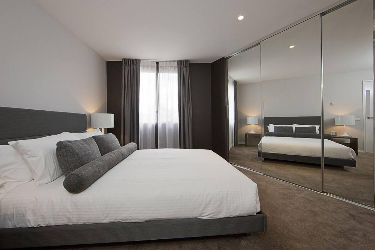 A modern hotel room with a large bed, a mirror wall, and a view of the adjacent room.