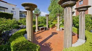 Une cour ornée de colonnes classiques, dotée d’un sol carrelé et bordée de haies parfaitement taillées.