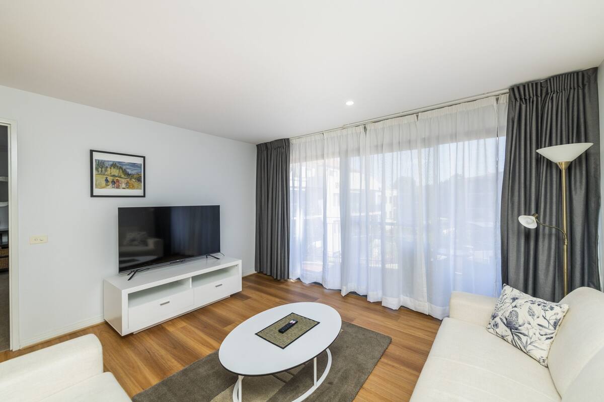 A modern living room with a flat-screen TV on a white TV stand, a white sofa with a patterned pillow, a small white coffee table, and a large window with grey curtains.