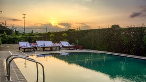 A swimming pool with lounge chairs and a netted area, surrounded by greenery. - Corbett Solitaire (Ramnagar)