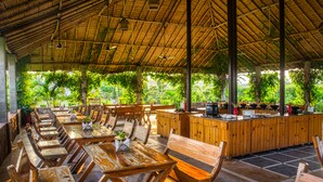 An outdoor dining area with wooden tables and chairs, a bar area, and a view of lush greenery. - Corbett Solitaire (Ramnagar)