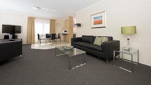 Ein modernes Hotelzimmer mit einem schwarzen Sofa, zwei Glastischen, einem Flachbildfernseher, einem Essbereich mit Tisch und Stühlen sowie einer Küchenzeile mit einer Mikrowelle.