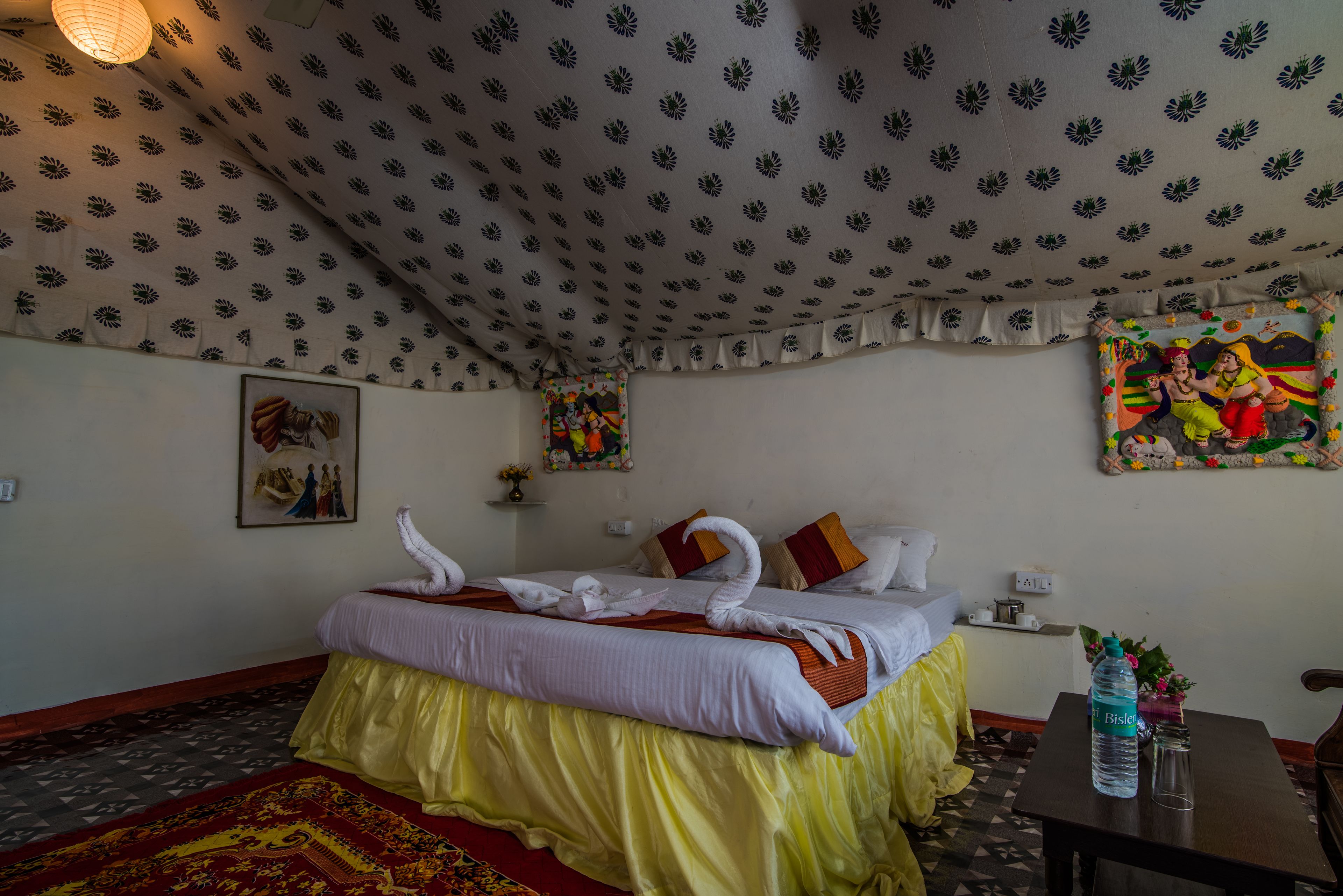 A hotel room with a patterned ceiling, a bed with decorative pillows, a water bottle on a table, and a colorful painting on the wall.