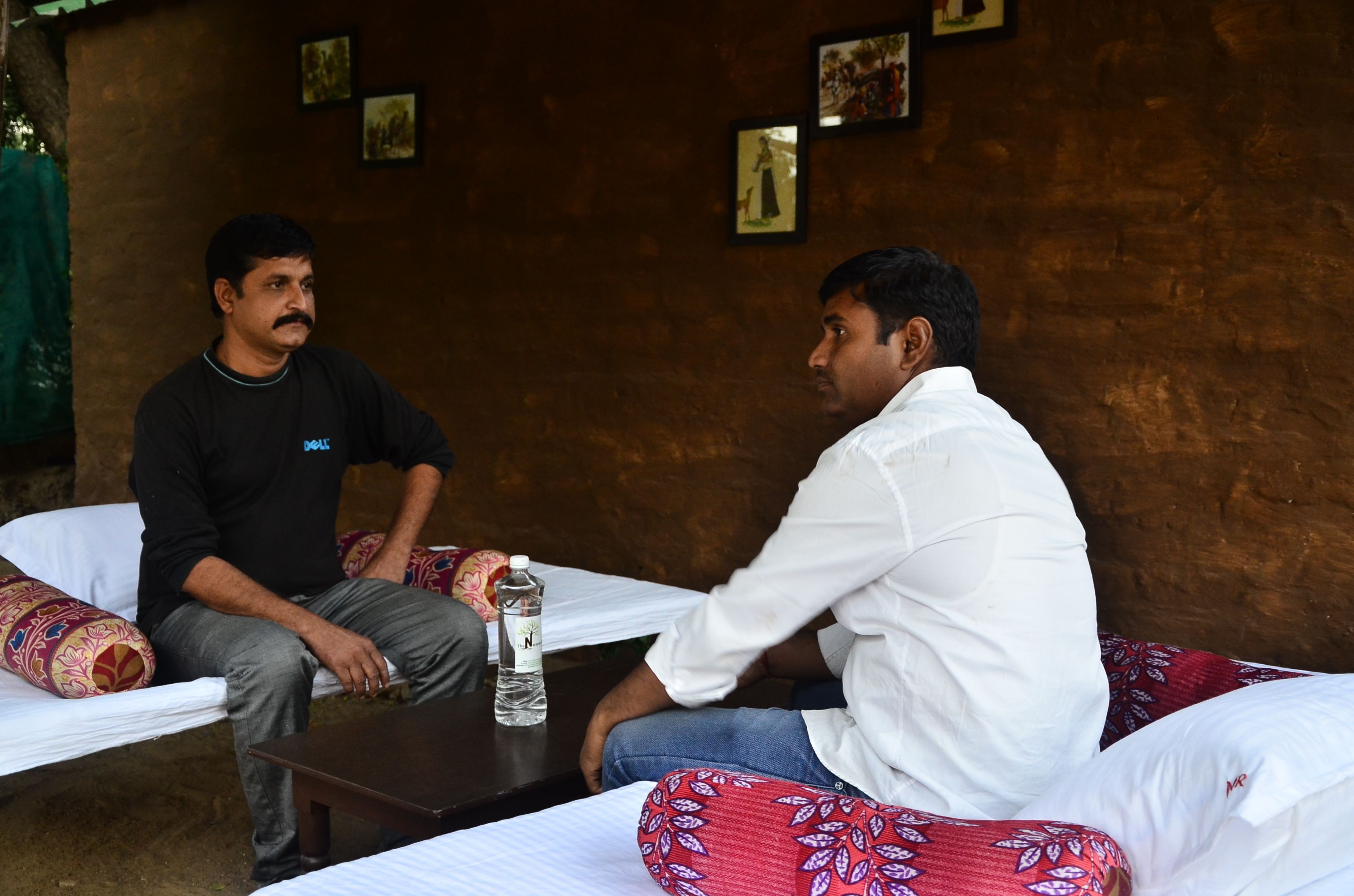 Two men seated on a bed with a water bottle on a table between them.