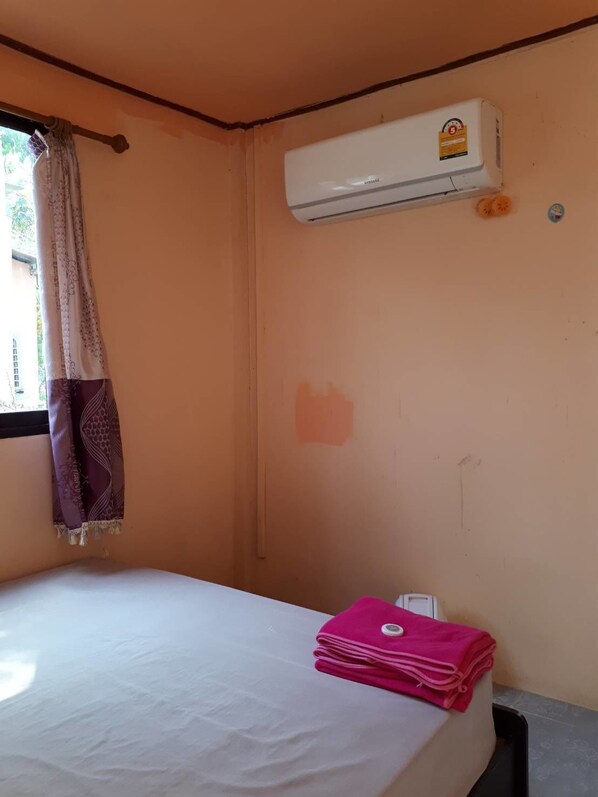 Une petite pièce avec un lit, une fenêtre avec un rideau, un climatiseur et une pile de serviettes.