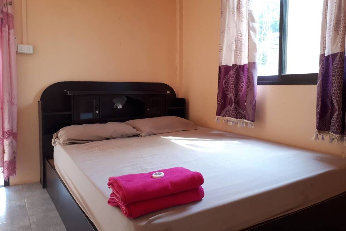 A single bed with a headboard, a window with curtains, and a wall socket.