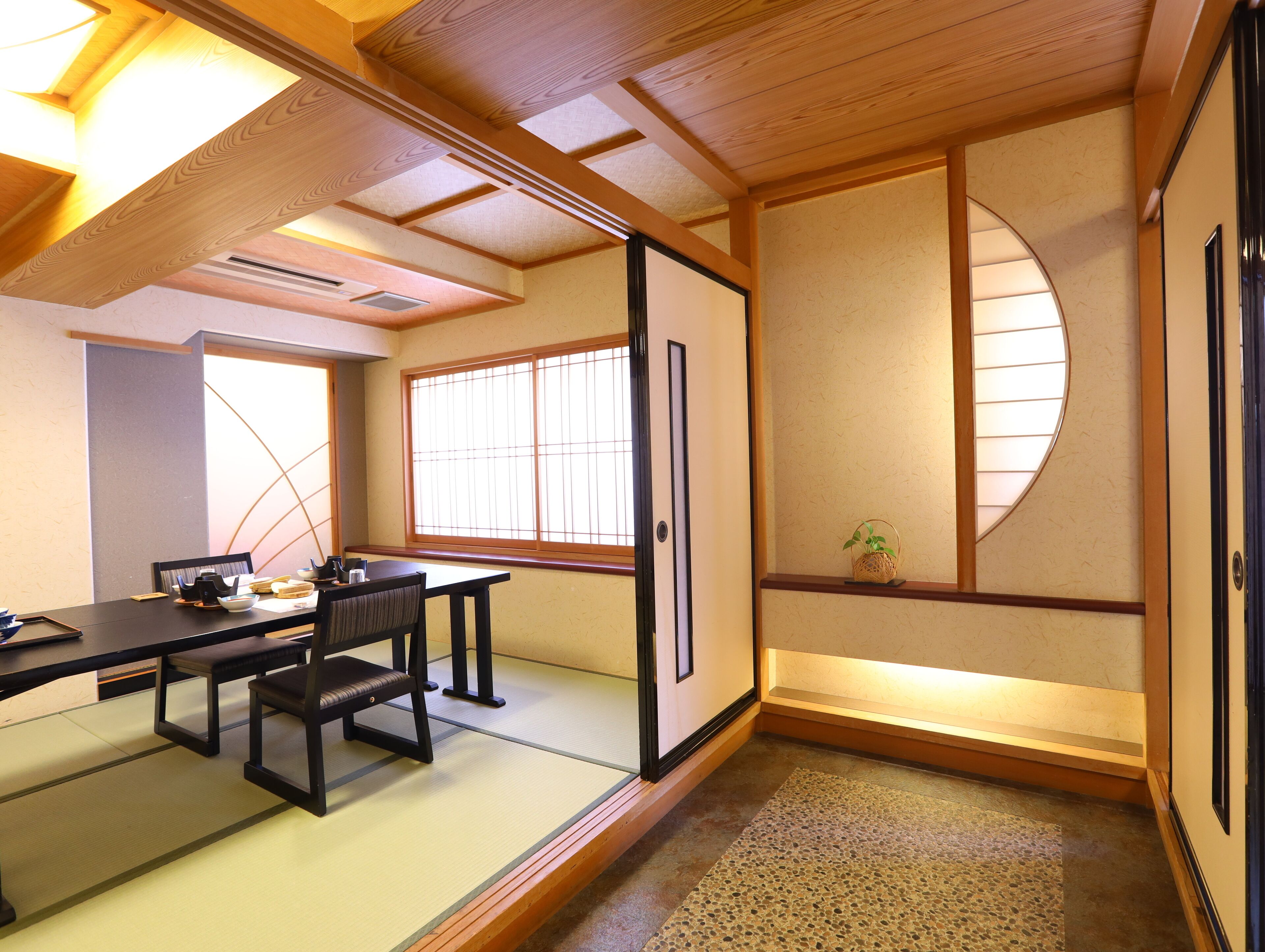A traditional Japanese room with tatami flooring, a low table with chairs, a sliding door, and a window with a view of the outside.
