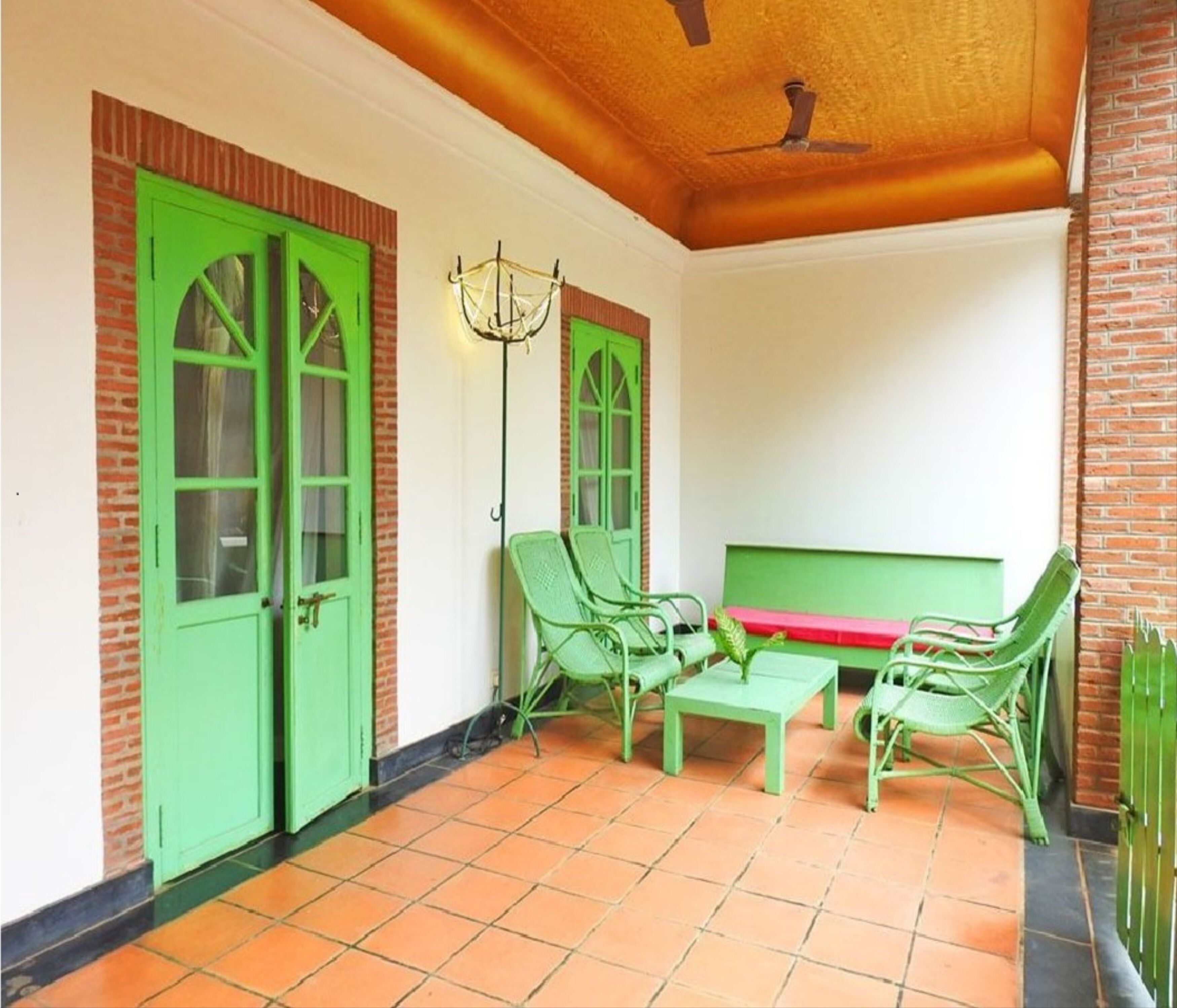 Een overdekt terras met groene stoelen en een tafel, bakstenen muren en een plafondventilator.