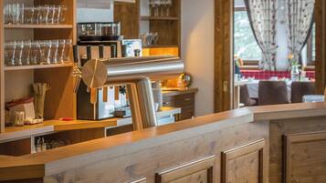 Zona de bar con barra de madera, estantes con vasos y utensilios de cocina, una caja registradora y vista al comedor a través de una ventana.