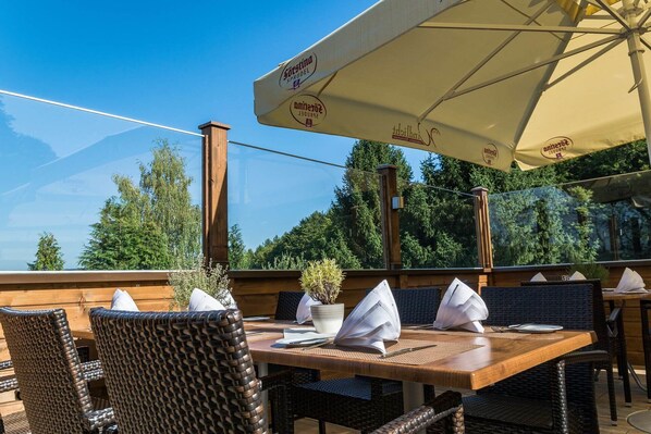 Un espacio de comedor al aire libre con mesas de madera y sillas de mimbre, grandes sombrillas y vistas a árboles y montañas.