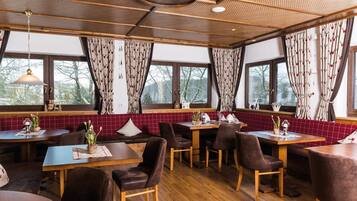 Interior de restaurante con mesas y sillas de madera, paredes con azulejos rojos y ventanas grandes con cortinas estampadas.