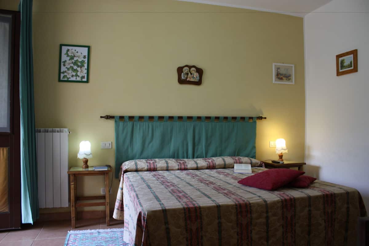 Quarto com cama, duas luminárias de cabeceira, mesa de cabeceira, radiador e janela com cortinas.