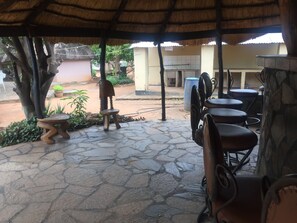 A covered outdoor seating area with stone flooring, a thatched roof, and a view of a building in the background. - Kana Guest House (Livingstone)