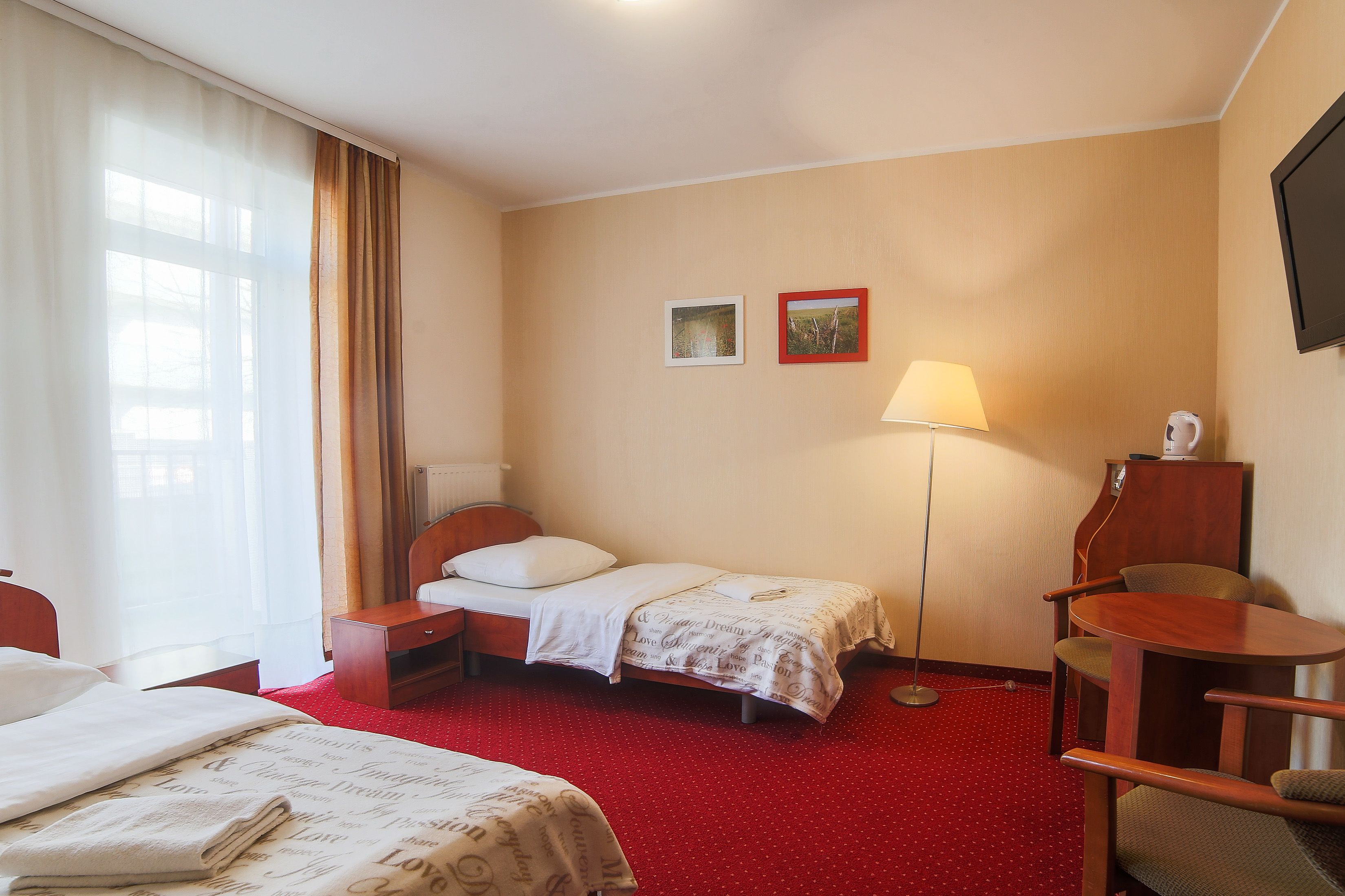 A hotel room with two single beds, a desk, a chair, a lamp, and two framed pictures on the wall.