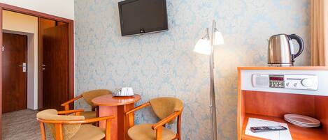 Habitación de hotel con televisor de pantalla plana montado en la pared, un escritorio de madera con una silla, una radio y una tetera sobre una estantería.