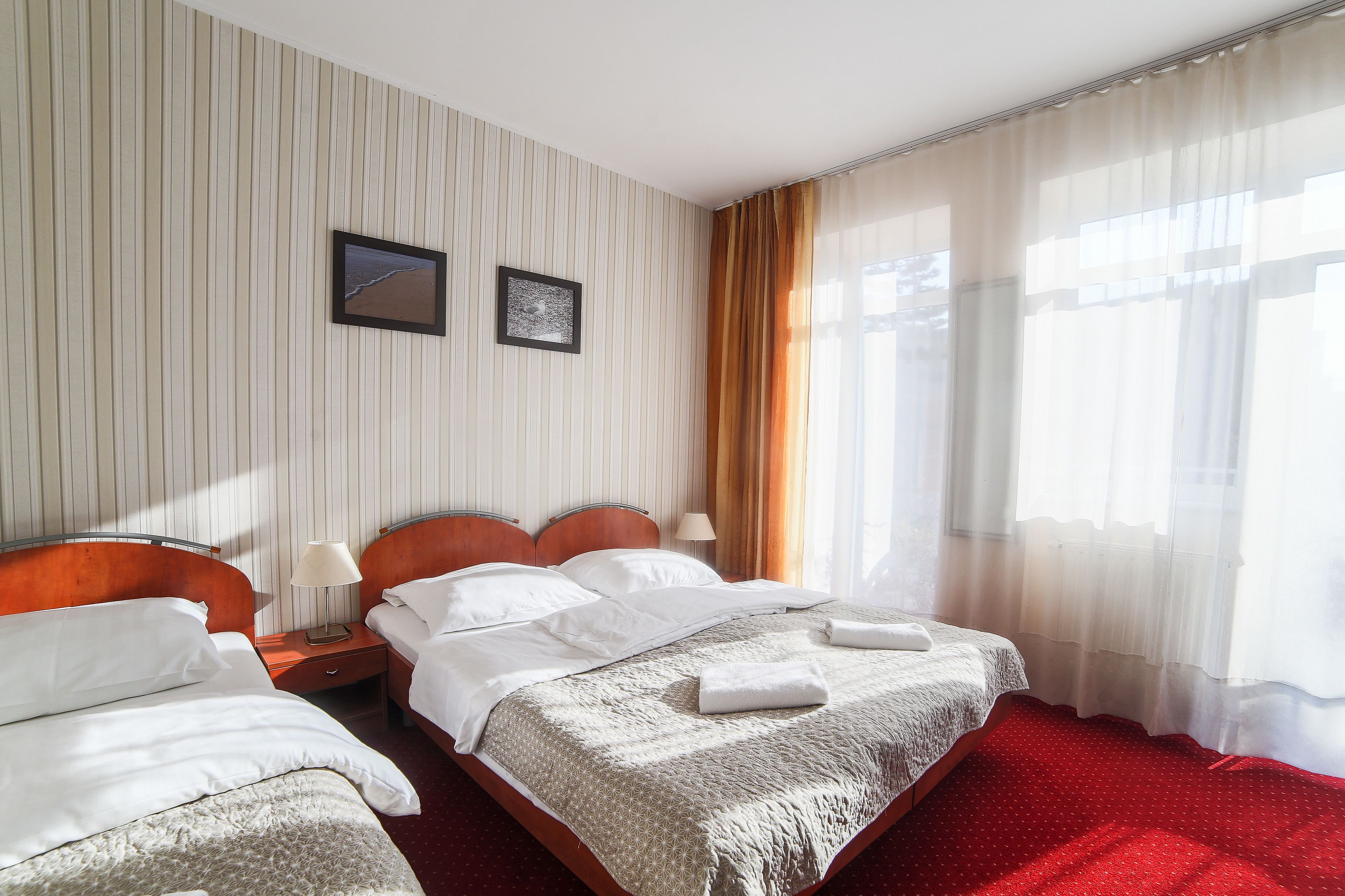 Une chambre d’hôtel avec deux lits, une moquette rouge, un papier peint à rayures et une fenêtre avec des rideaux légers.