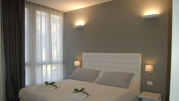 A neatly made bed with pillows and folded towels, a bedside table, and two wall-mounted lamps. - Midnight in Genova (Genoa)