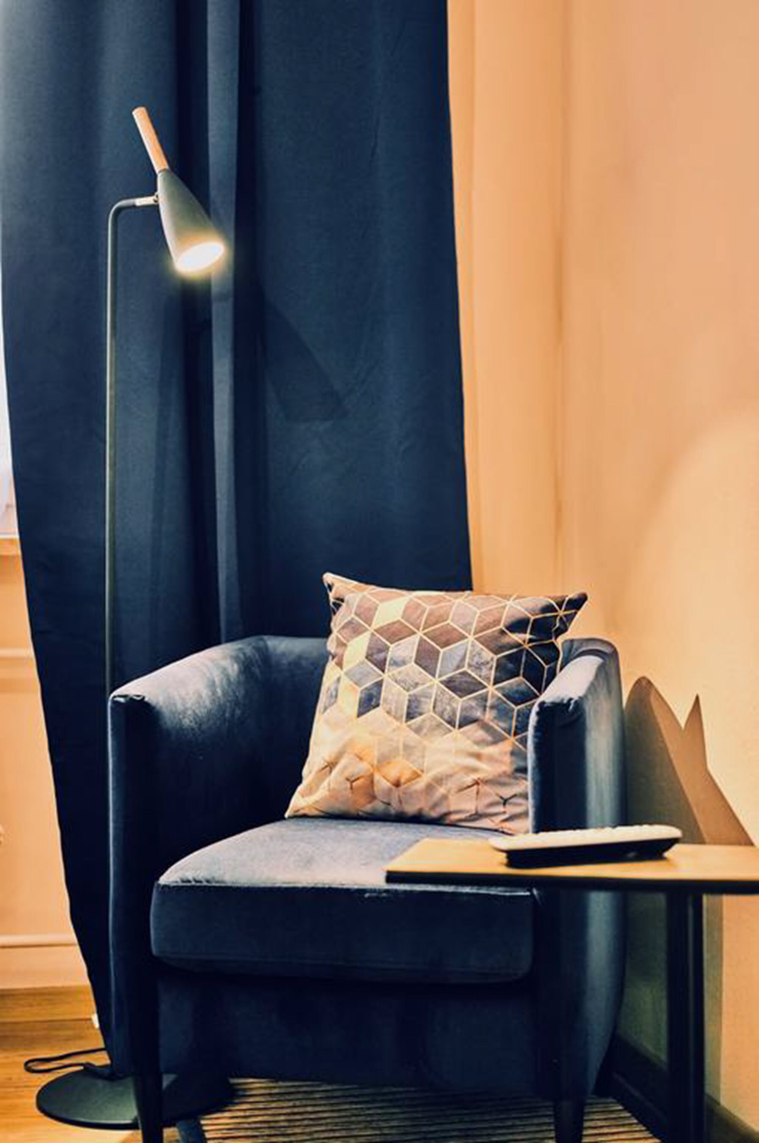 A modern room with a dark blue armchair, a geometric patterned pillow, a small wooden side table, and a floor lamp.