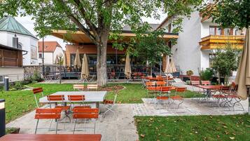 Un espace extérieur aménagé en terrasse, avec des tables et des chaises, entouré de verdure et bordé d’un bâtiment doté d’un balcon.