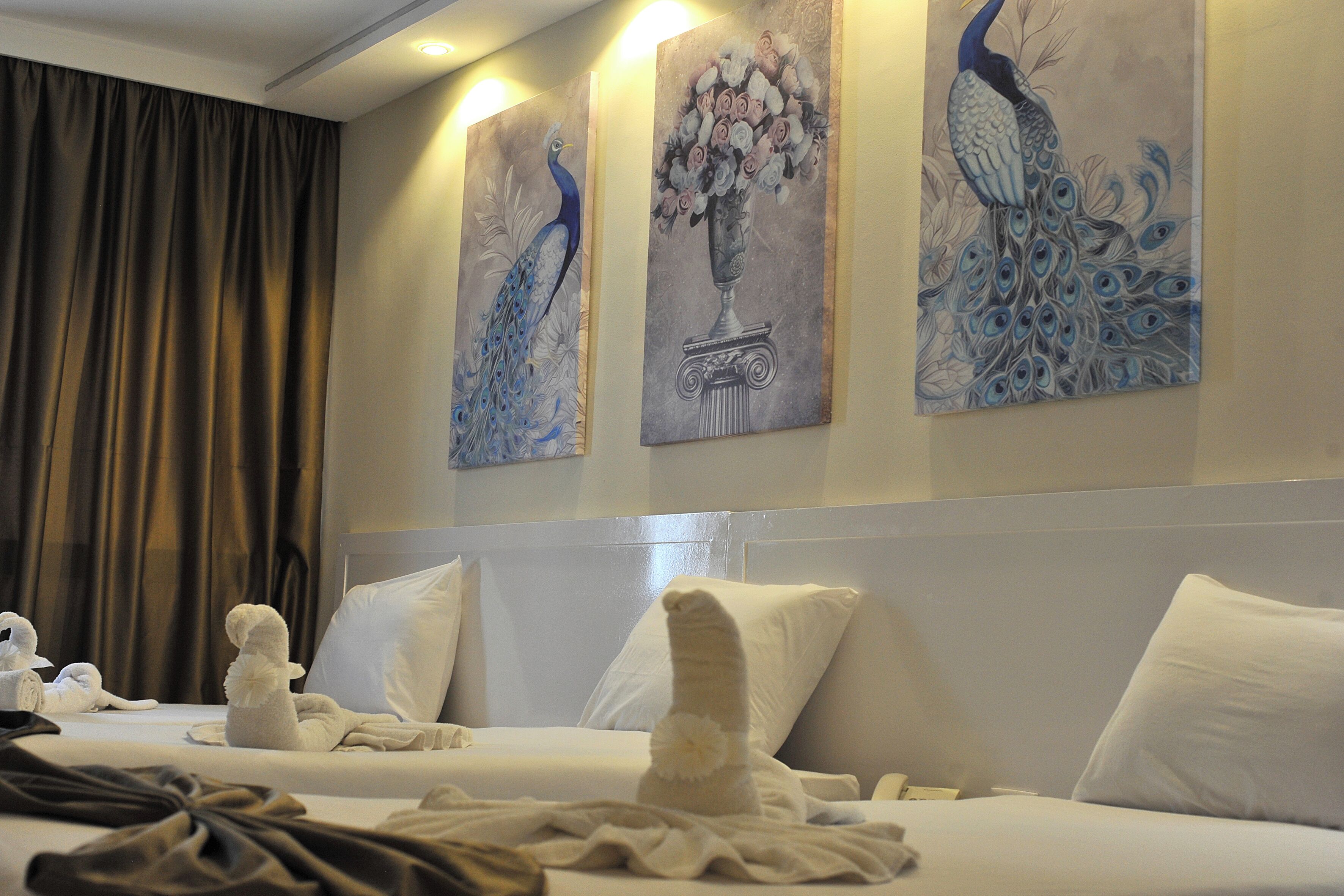 A hotel room with a bed, pillows, a headboard, and three wall-mounted paintings of peacocks.