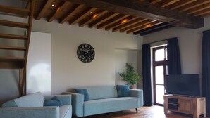 Een woonkamer met een houten plafond, een blauwe bank, een houten tv-meubel en een klok aan de muur.