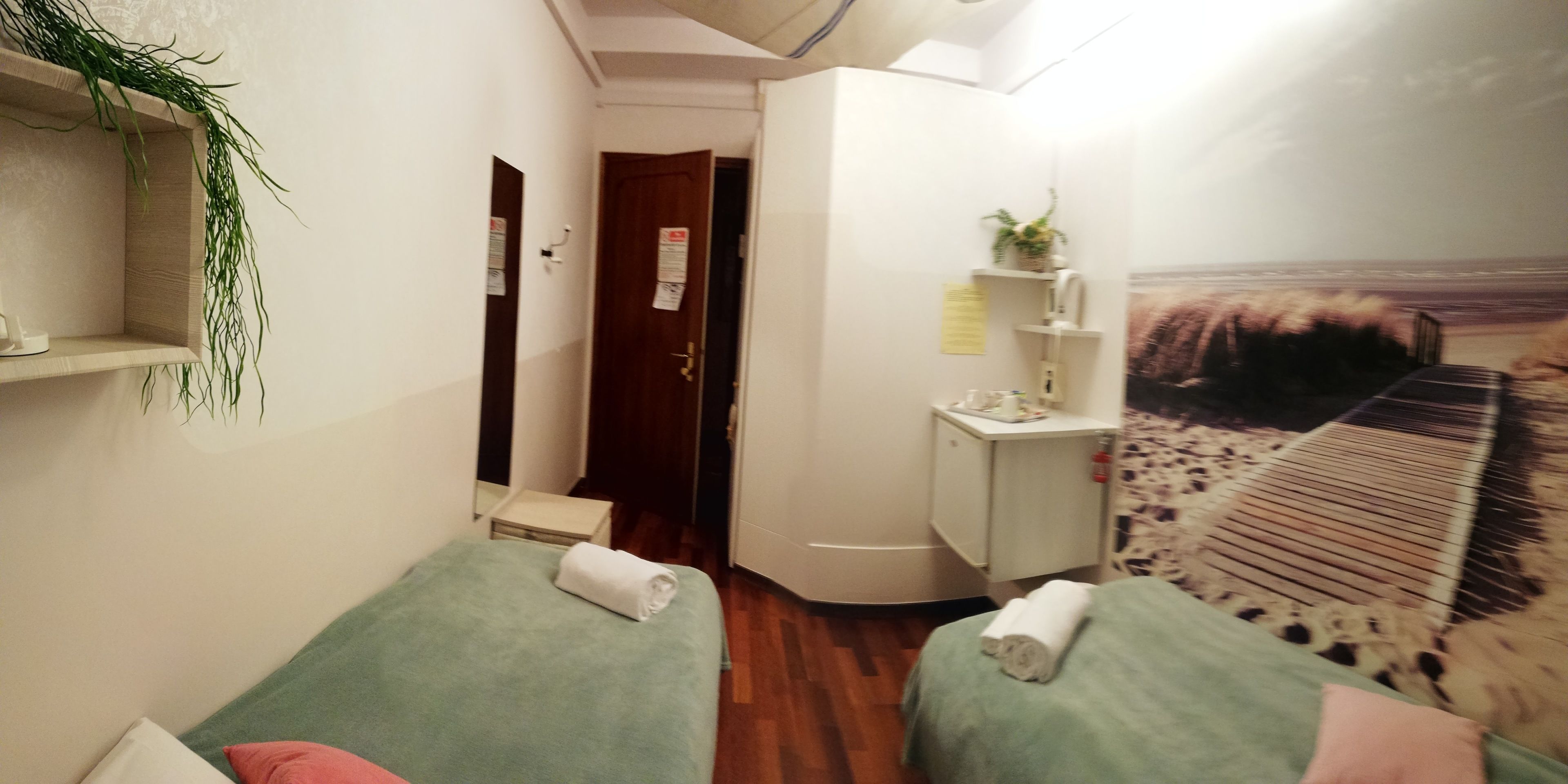 A hotel room with two beds, a small desk, a mirror, and a wall-mounted shelf with a plant.