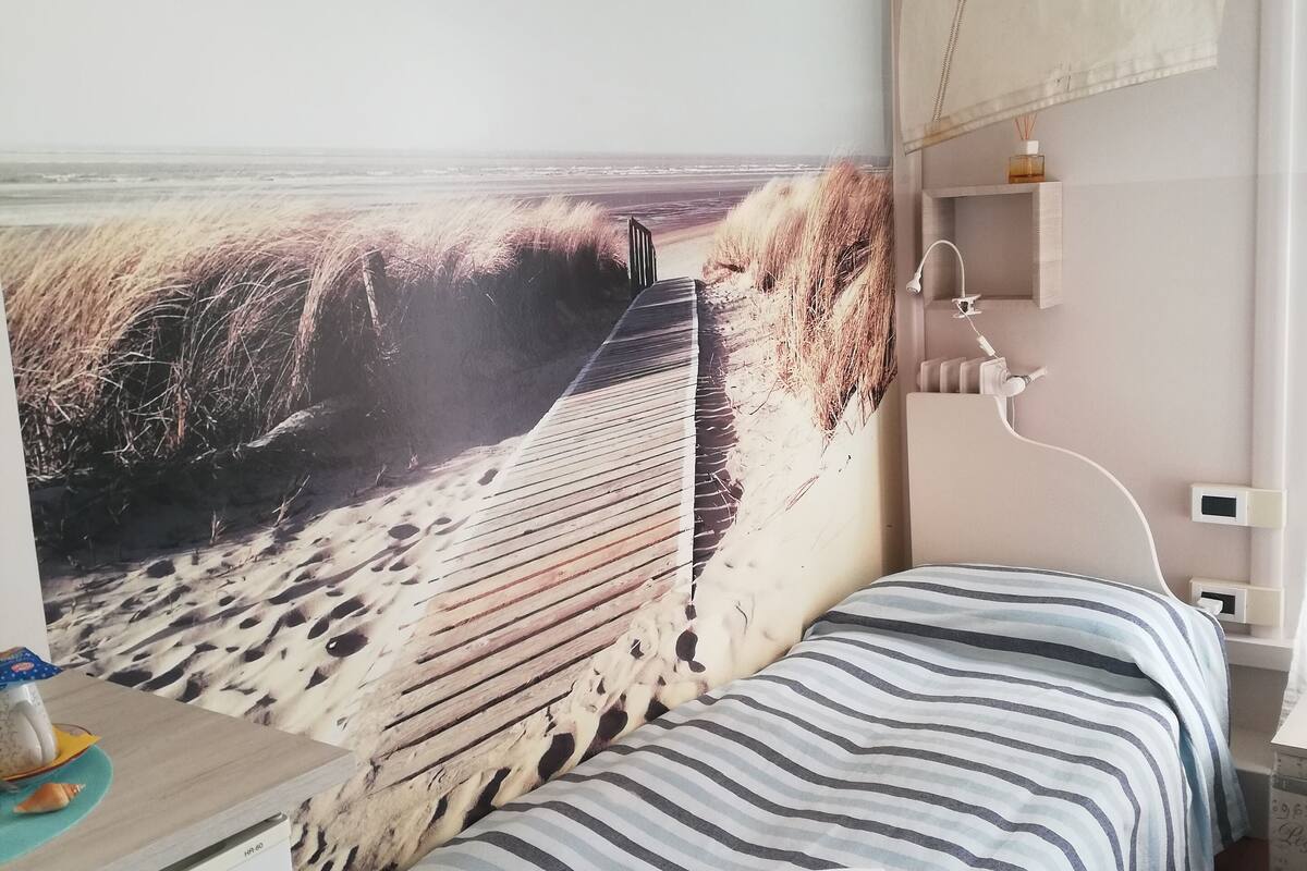 Un dormitorio con un mural de playa, una cama con ropa de cama a rayas, un escritorio y una mesita de noche pequeña.