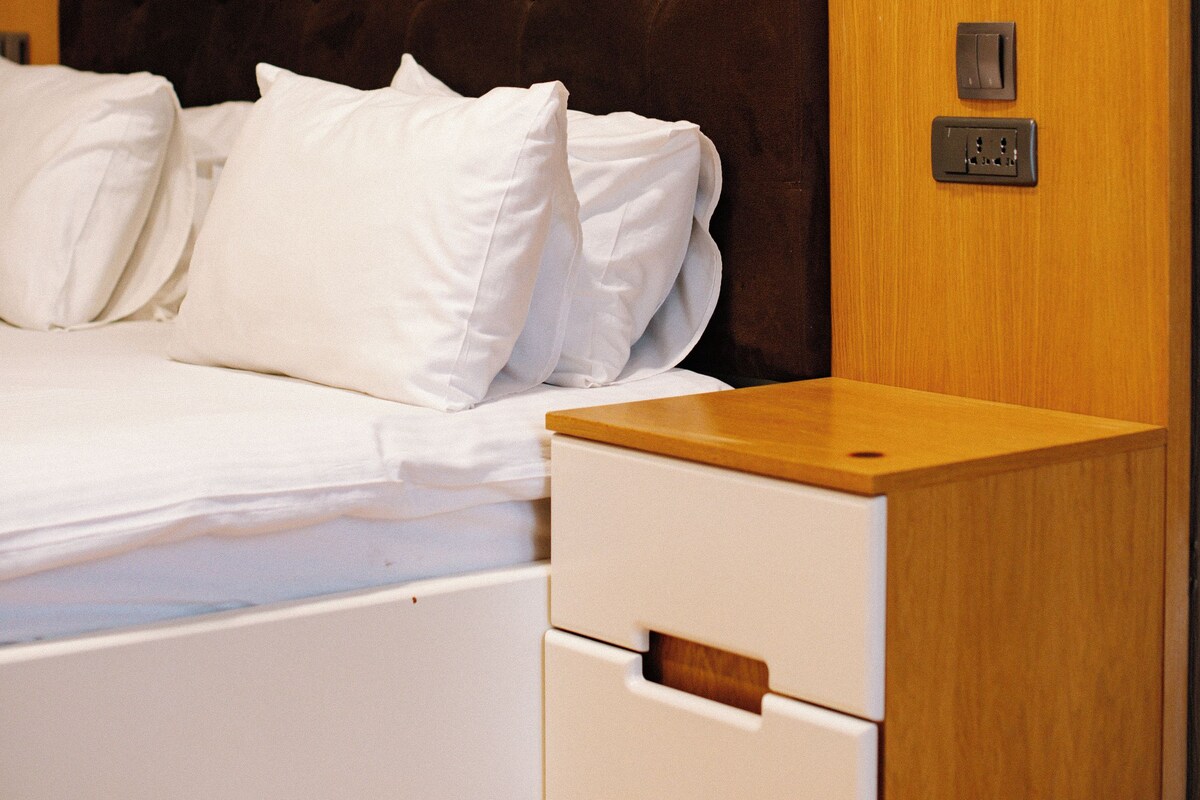 A neatly made bed with white linens and a wooden headboard. There is a wooden nightstand with a drawer and a wall socket.