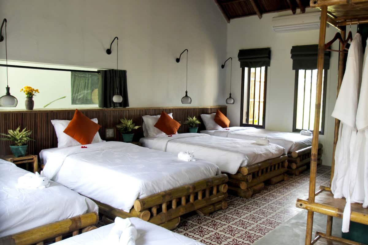 A hotel room with four wooden beds, each with white linens and orange pillows. There are potted plants on the nightstands, a mirror above the beds, and a wooden wardrobe with a white towel hanging on it.