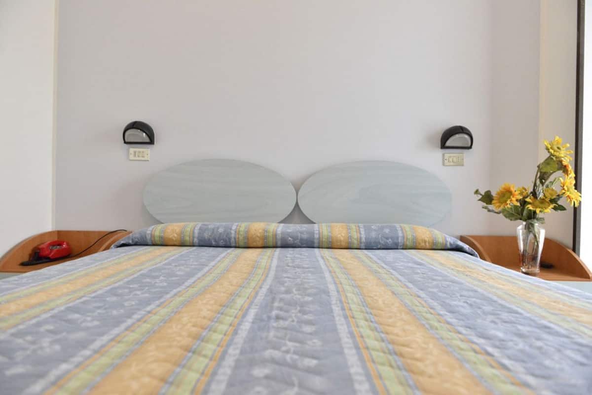 Uma cama com colcha listrada, duas mesas de cabeceira com abajures e um vaso com flores do lado direito.