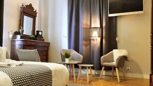 Et hotelværelse med en seng, to lænestole, et lille bord med en plante, en lampe, en kommode med et ur og et tv på væggen.