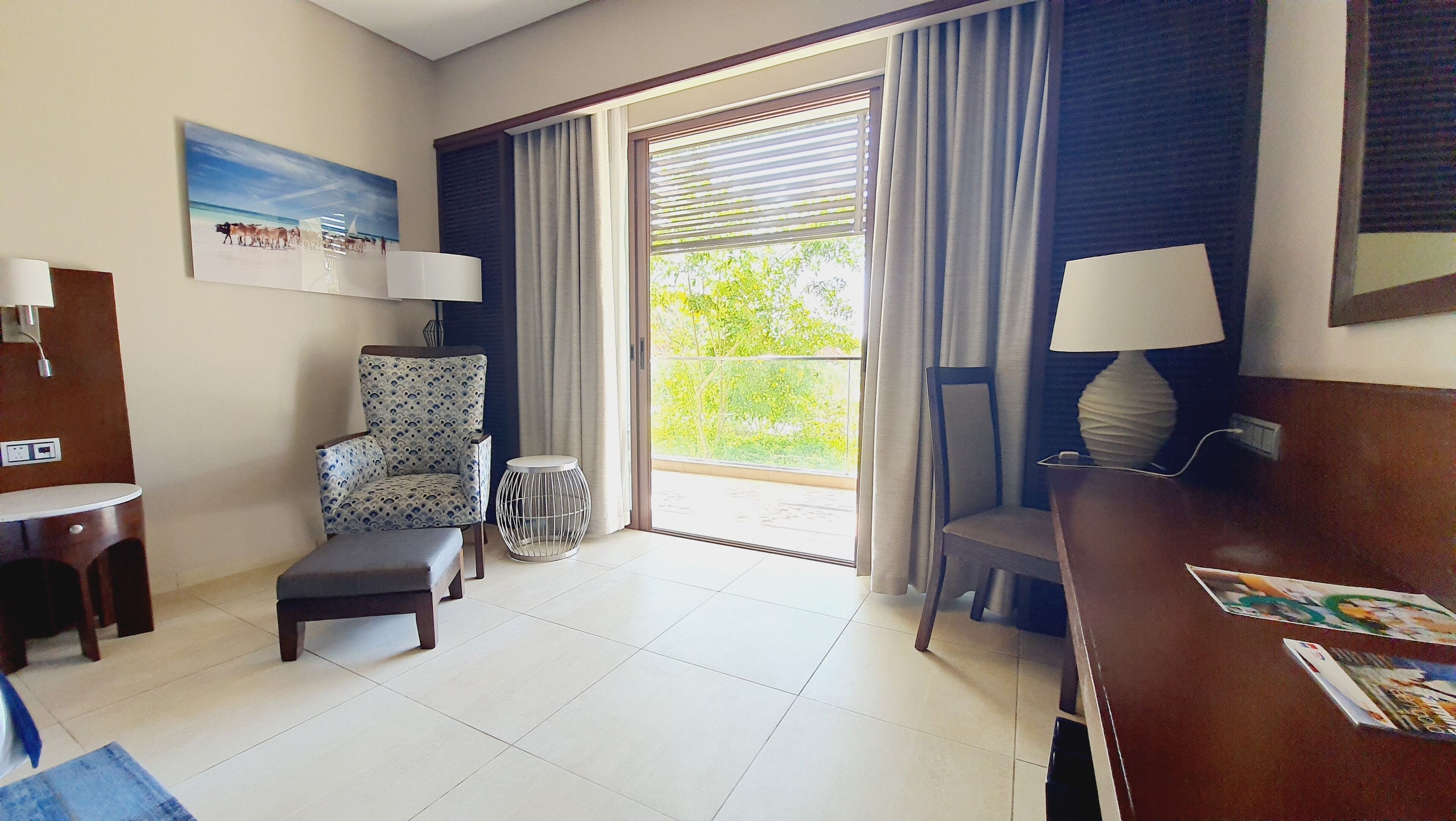 A hotel room with a large window, a chair, a small table, and a desk.