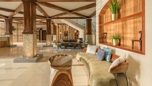 Un lobby amplio con techo alto, vigas de madera, un área de estar central con sofás y una mesa de centro, y un estante de madera con plantas.