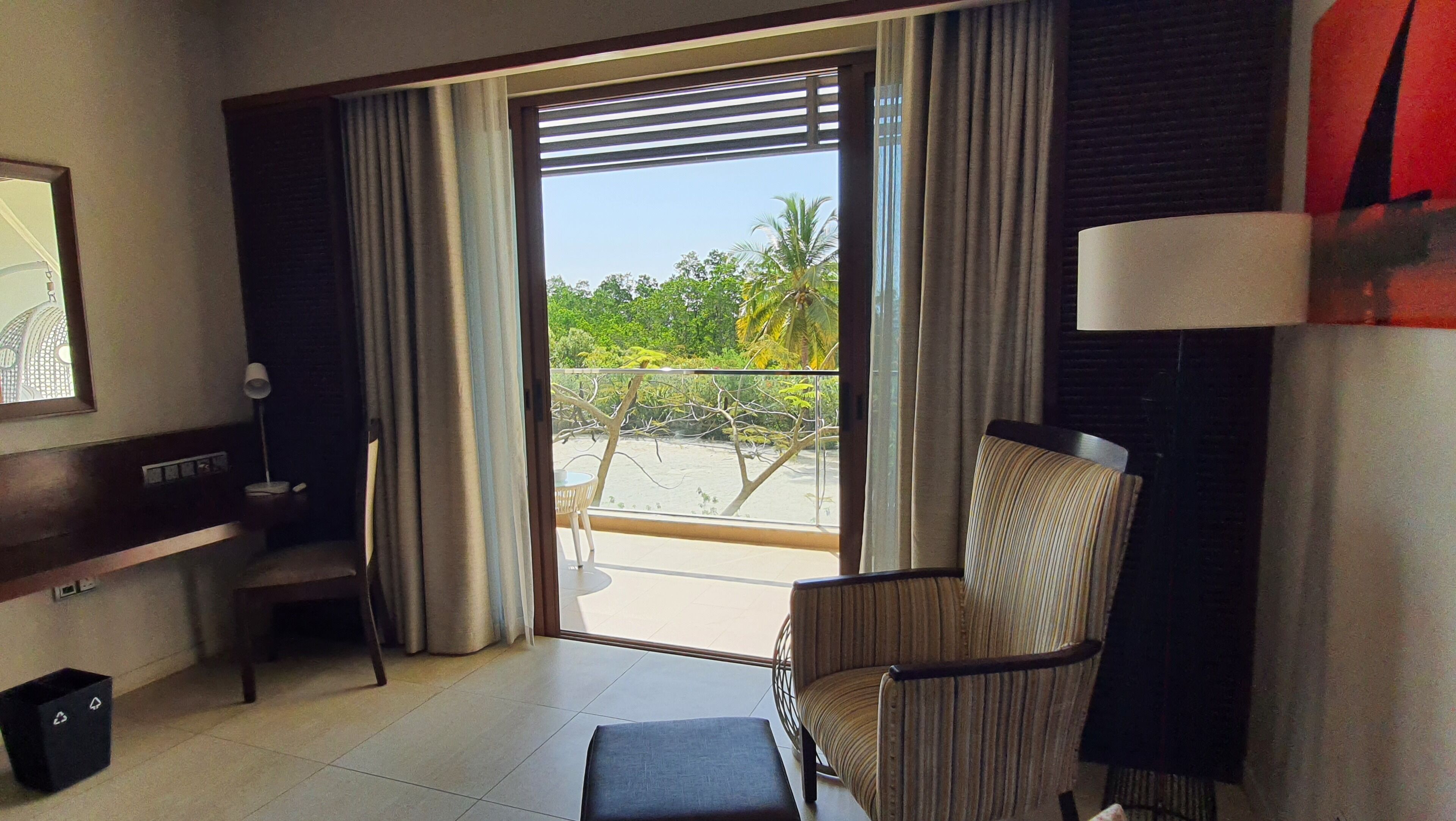 Quarto de hotel com varanda, uma cadeira, uma escrivaninha e um abajur.