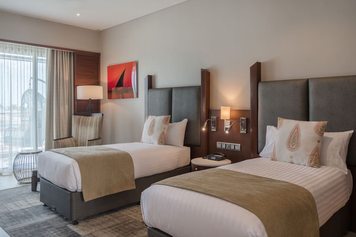 Ein Hotelzimmer mit zwei Betten, einem großen Fenster mit Vorhängen, einem Wandbild und einem kleinen Tisch mit einer Lampe.