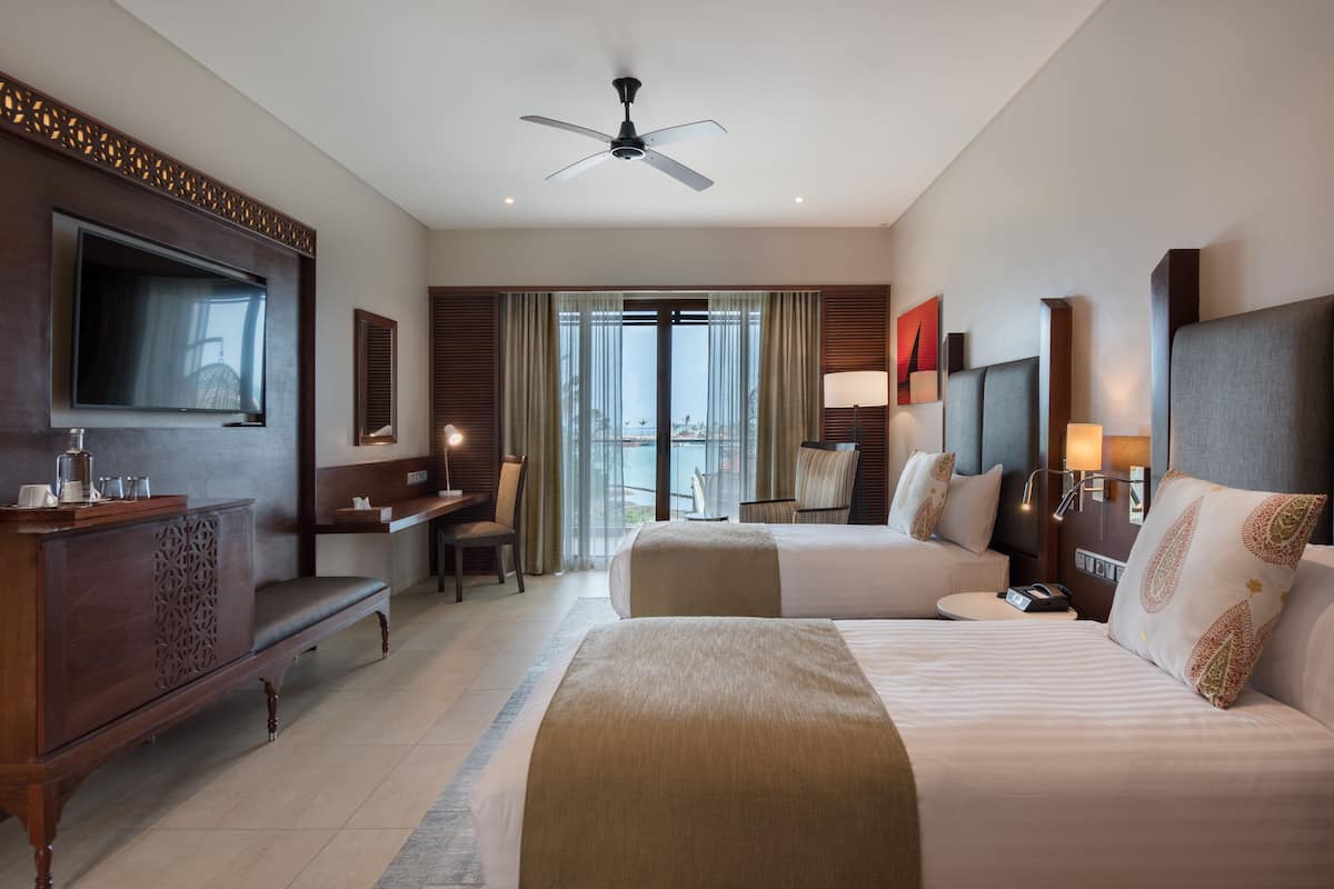 Ein Hotelzimmer mit zwei Betten, einem Fernseher, einem Schreibtisch und einem Balkon mit Vorhängen.