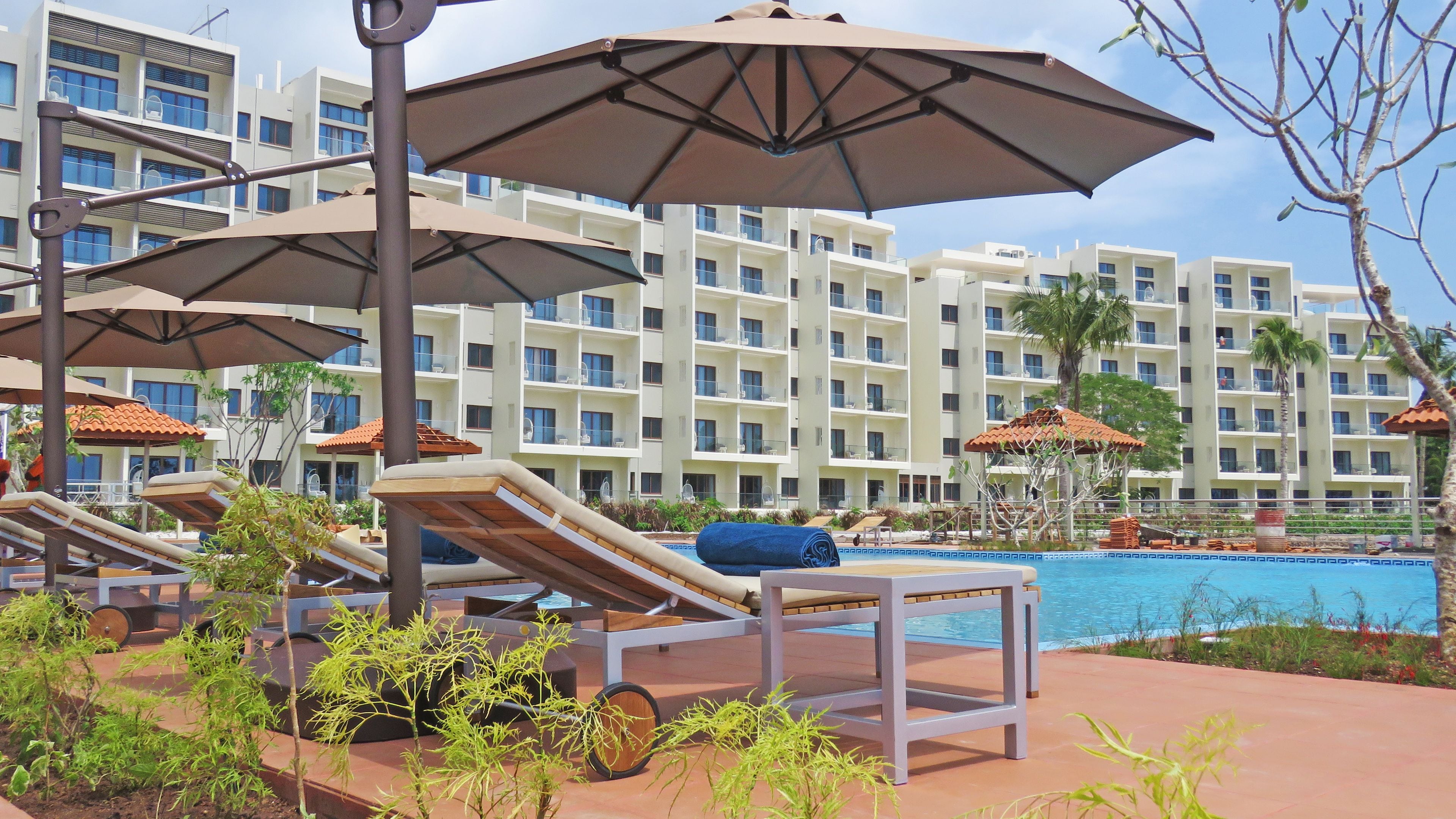 A poolside area with lounge chairs and umbrellas, a swimming pool, and a multi-story hotel building in the background.