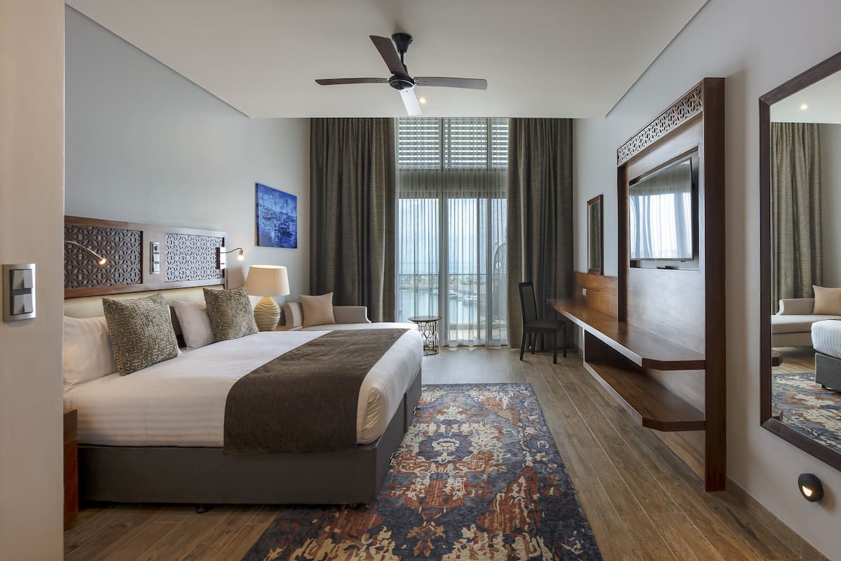 Ein Hotelzimmer mit einem großen Bett, einem Schreibtisch, einem Sessel und einem Deckenventilator, mit Blick ins Freie.