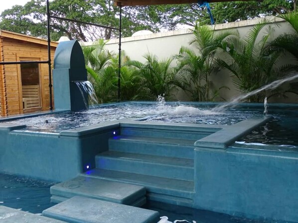 Un bain à remous avec des marches et une cascade, le tout entouré de verdure.