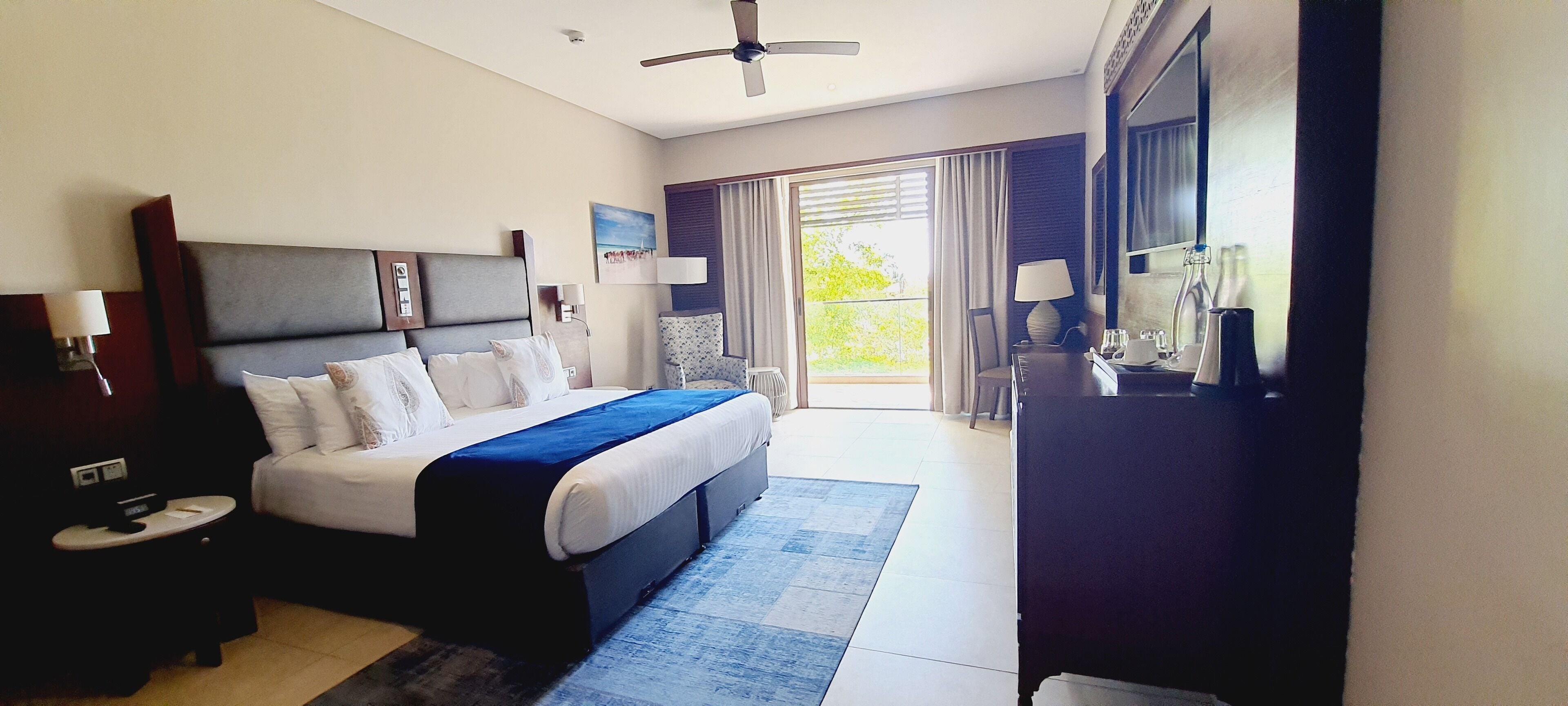 Habitación de hotel con una cama grande, un escritorio, una silla, un televisor y un balcón con cortinas.