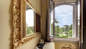 Ein Zimmer mit einem großen, verzierten Spiegel, einem Fenster mit Blick auf Grünflächen und einem Tisch mit einem Tablett mit Gläsern und einer Tasse.