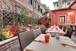 An outdoor dining area with brick walls, a lattice trellis, and a variety of colorful flowers. Tables are set with wine glasses, cutlery, and napkins.