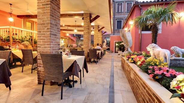 Ein Essbereich im Freien mit Ziegelstützen, Tischen mit weißen Tischdecken und einem dekorativen Pflanzgefäß mit Blumen und Statuen.