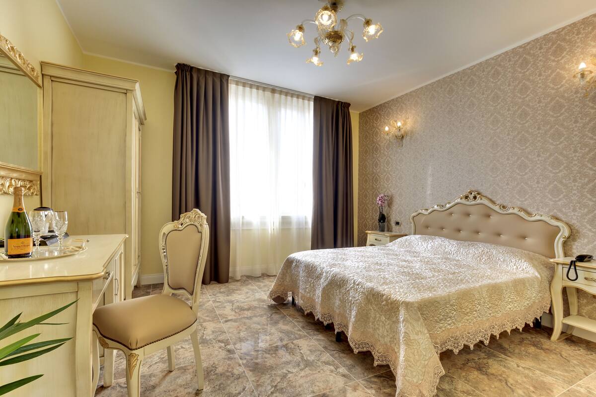 A bedroom with a large bed, a desk with a chair, a chandelier, and a window with curtains.