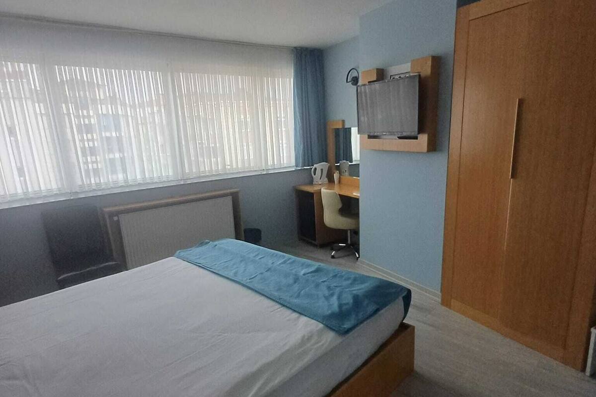 Ein Hotelzimmer mit einem Bett, einem Schreibtisch, einem Stuhl, einem Kleiderschrank, einem Fenster mit Jalousien und einer Deckenleuchte.