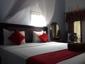 A bedroom with a bed, red and white pillows, a bedside table with a lamp, a mirror, and a window with curtains. - Mangga Homestay (Gili Air)