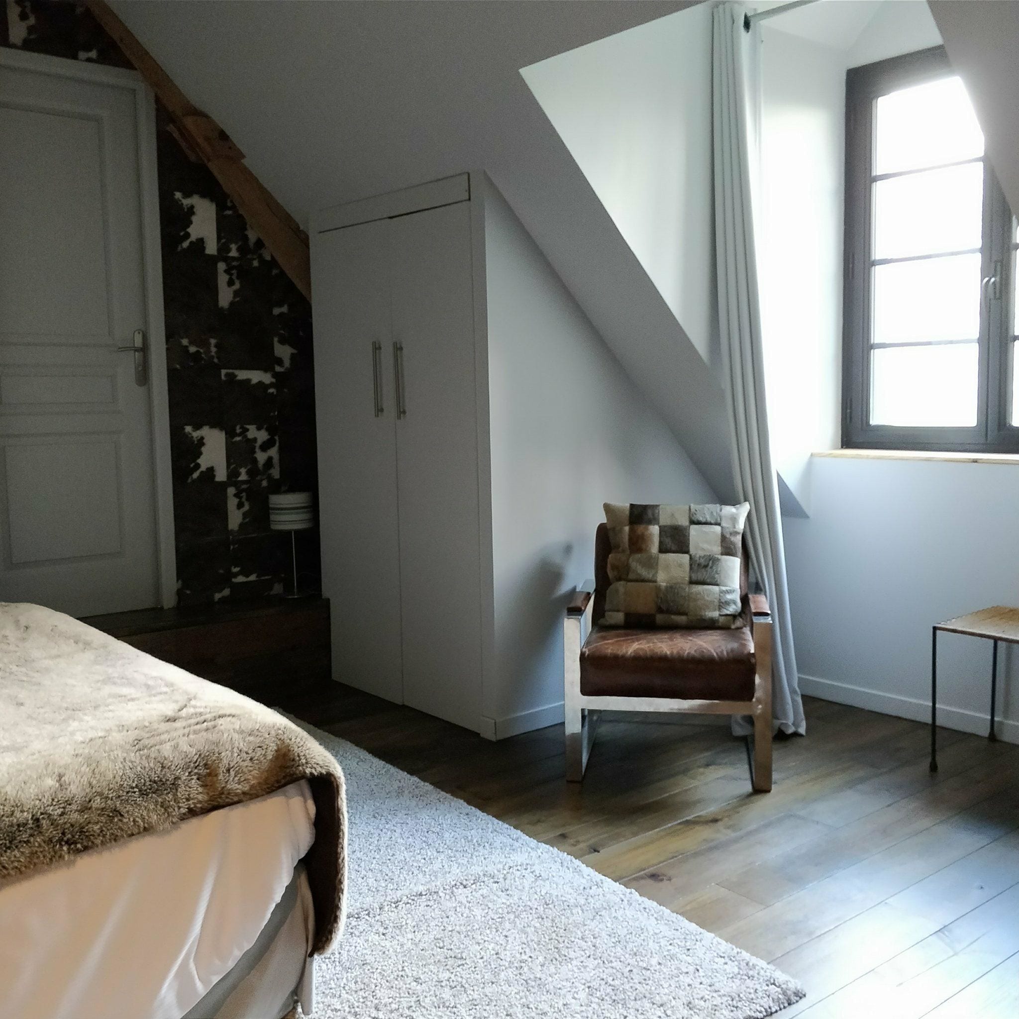 A bedroom with a sloped ceiling, a bed, a chair with a patterned cushion, a wardrobe, and a window with curtains.