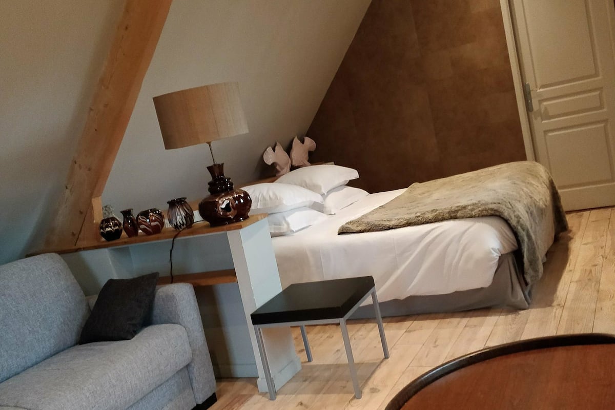 A cozy room with a sloped ceiling, a bed with white bedding, a grey sofa, and a small table with decorative items.