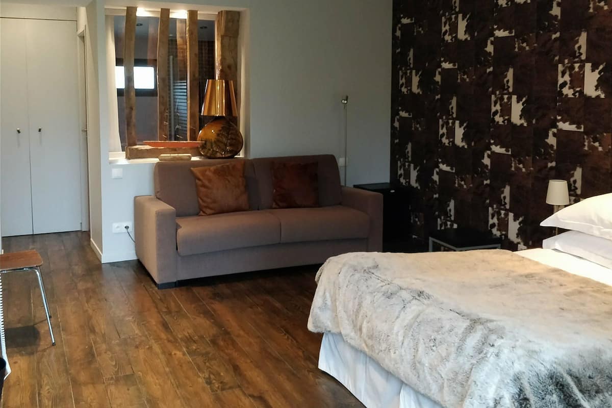A bedroom with a bed, a sofa, a wooden ceiling beam, a mirror, and a patterned wall.
